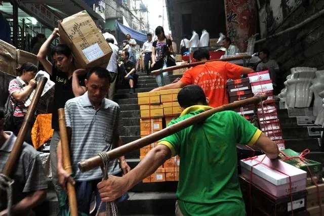 An elderly porter carrying a heavy load of cardboard boxes balanced on a green bamboo pole over his shoulder up concrete stairs.