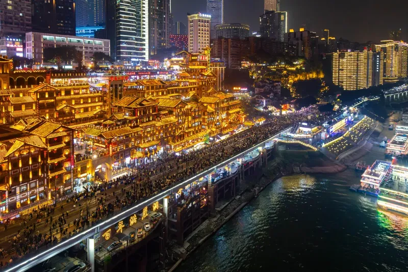 A towering wooden structure clinging to a steep cliff, brightly lit with golden neon lights against the dark night sky next to a river.