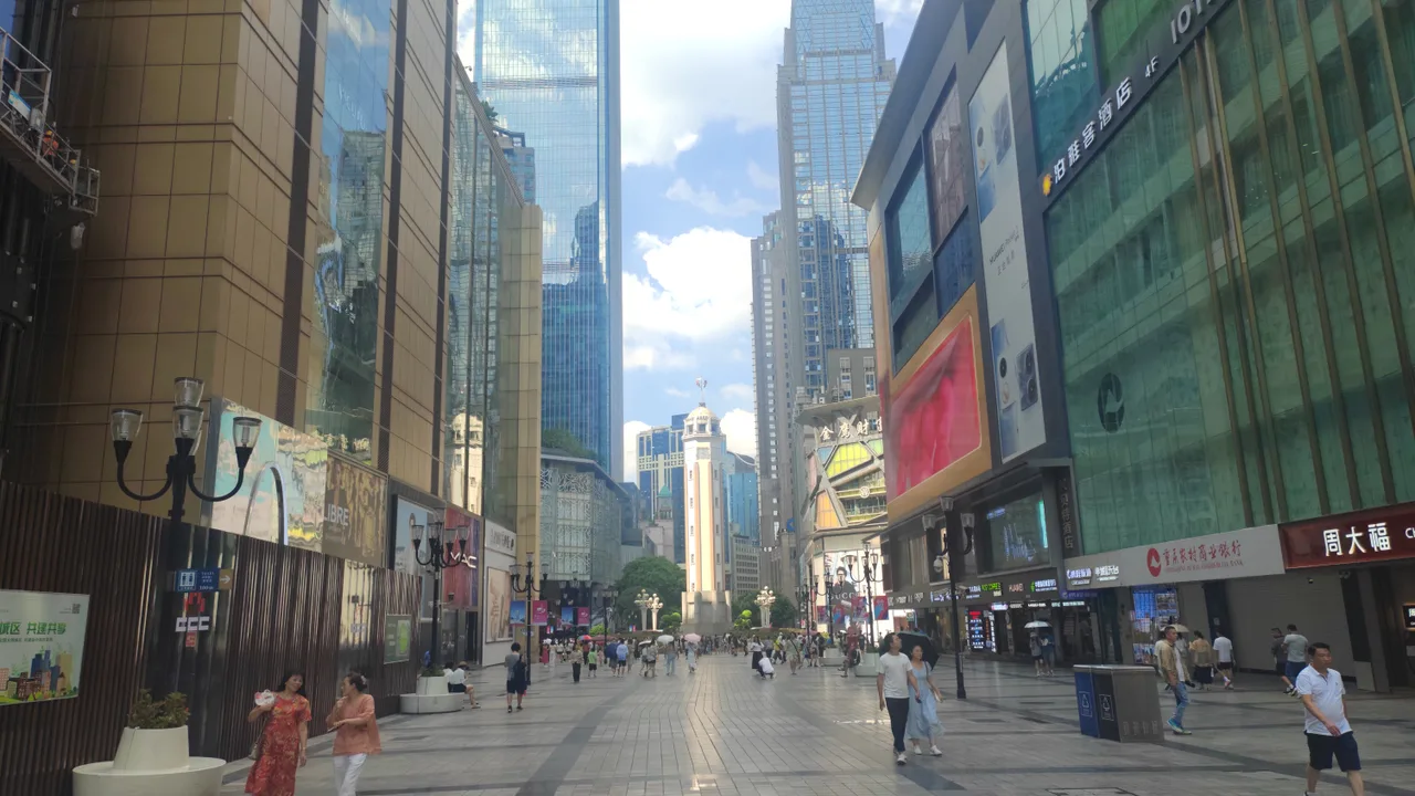 The Jiefangbei pedestrian street in Chongqing on a sunny day, with glass towers on both sides and the Liberation Monument clock tower at the far end.