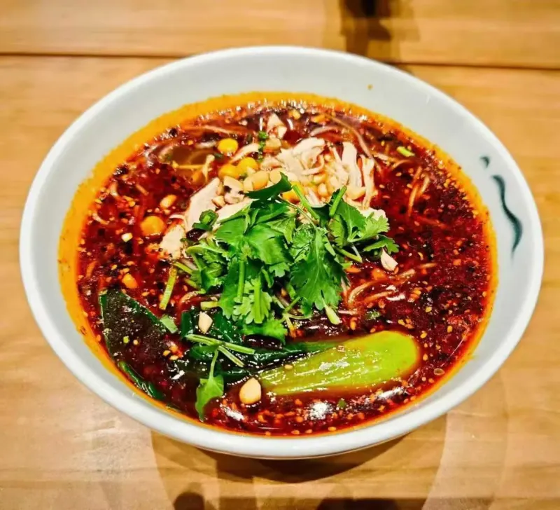 A steaming bowl of noodles topped with slow-cooked yellow peas, minced pork, and bright red chili oil on a small plastic stool.