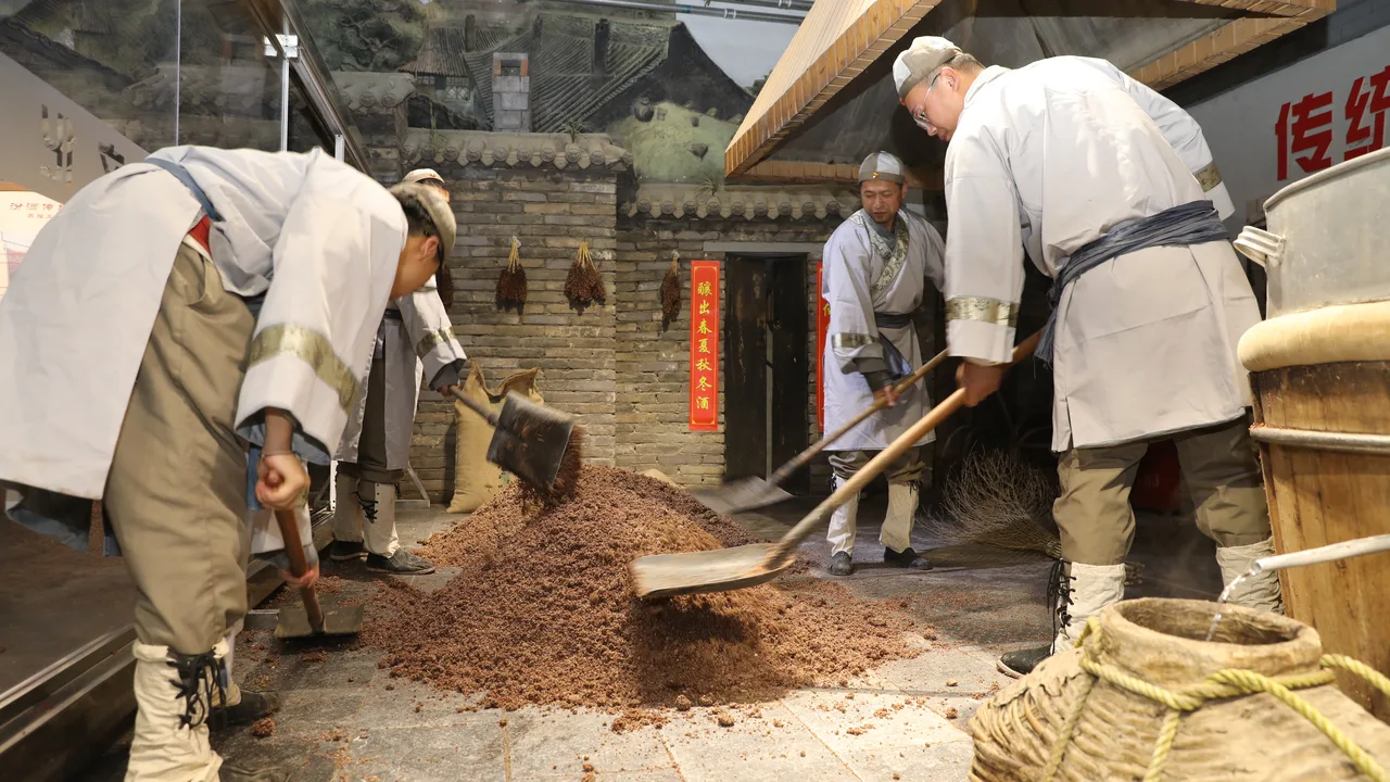 Workers in traditional white uniforms shovelling steamed grain at the reconstructed old production line, Fenjiu Culture Scenic Area, Xinghuacun.