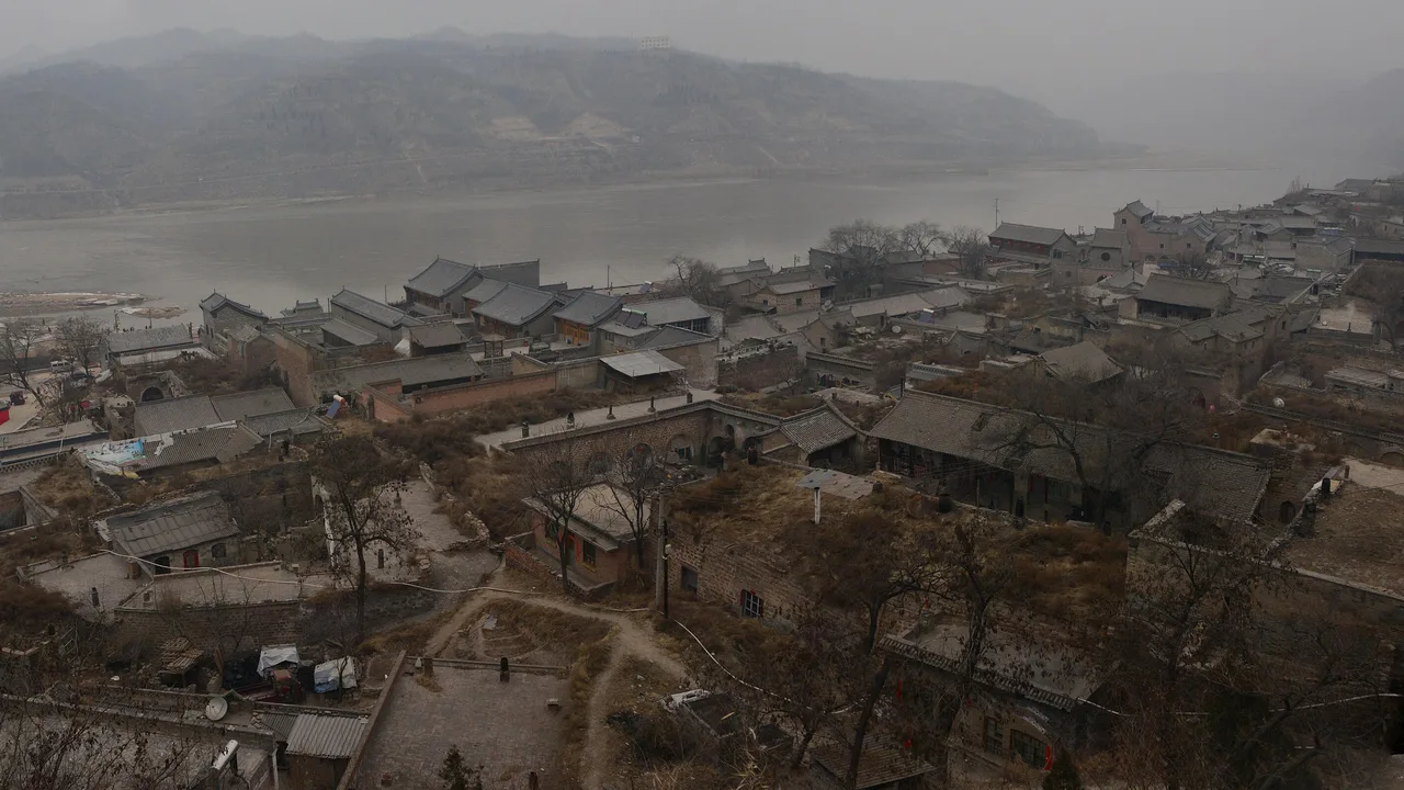 Overview of Qikou's old town rooftops on the slope above the Yellow River, with the Shaanxi hills on the far bank under hazy sky.