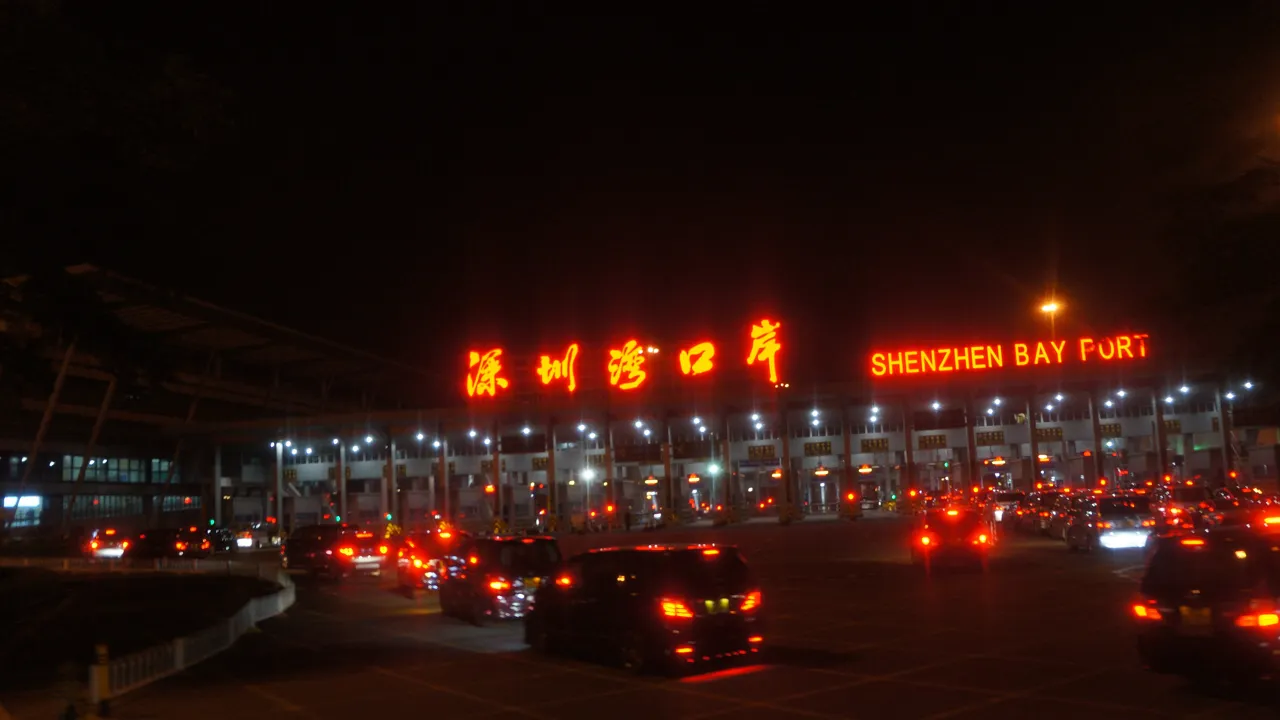 Shenzhen Bay Port at night, its red neon sign in Chinese and English glowing above the vehicle clearance lanes as cars queue toward the booths.