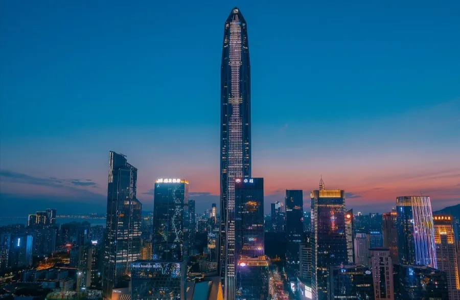 The towering glass spire of the Ping An Finance Centre rising high above the surrounding skyline and street-level parks in Futian, Shenzhen.