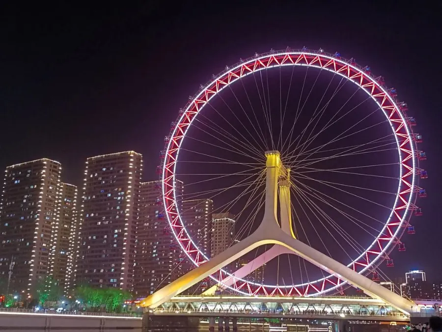 The illuminated Tianjin Eye Ferris wheel standing tall over the Yongle Bridge at night, reflecting in the Haihe River.