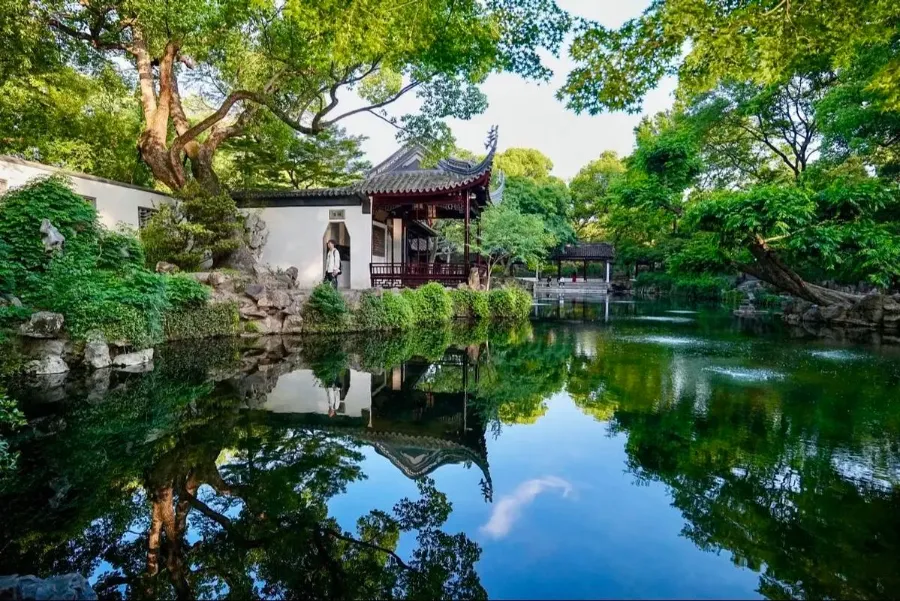 A stone bridge crosses the green waters of Jichang Garden's central pond, with traditional pavilions and leafy trees.