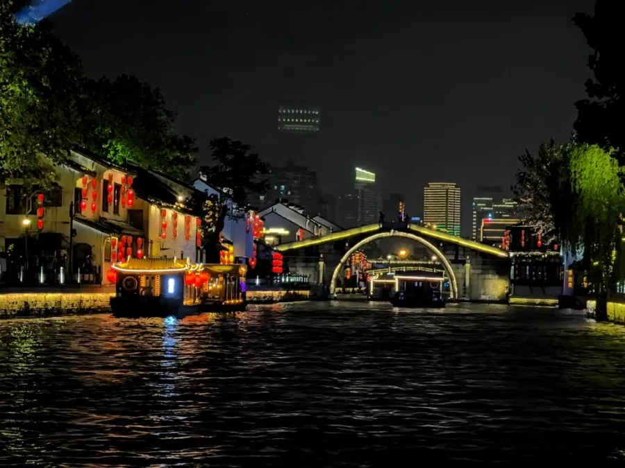 A stone arch bridge lit by red lanterns reflecting in the dark waters of the Grand Canal at night.