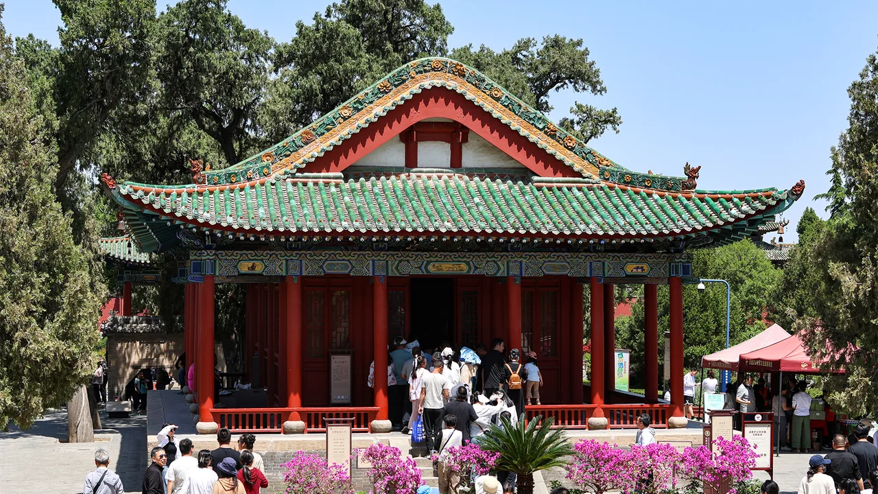 Red-pillared gateway pavilion with green-glazed roof at Xiezhou Guandi Temple, Yuncheng, with visitors and flower beds in front.